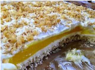 Μια εντυπωσιακή τούρτα λεμονιού σε στρώσεις, με λευκή κρέμα τυριού, κίτρινη λεμονόκρεμα και αφράτη σαντιγί, πασπαλισμένη με λευκή σοκολάτα και καρύδια.