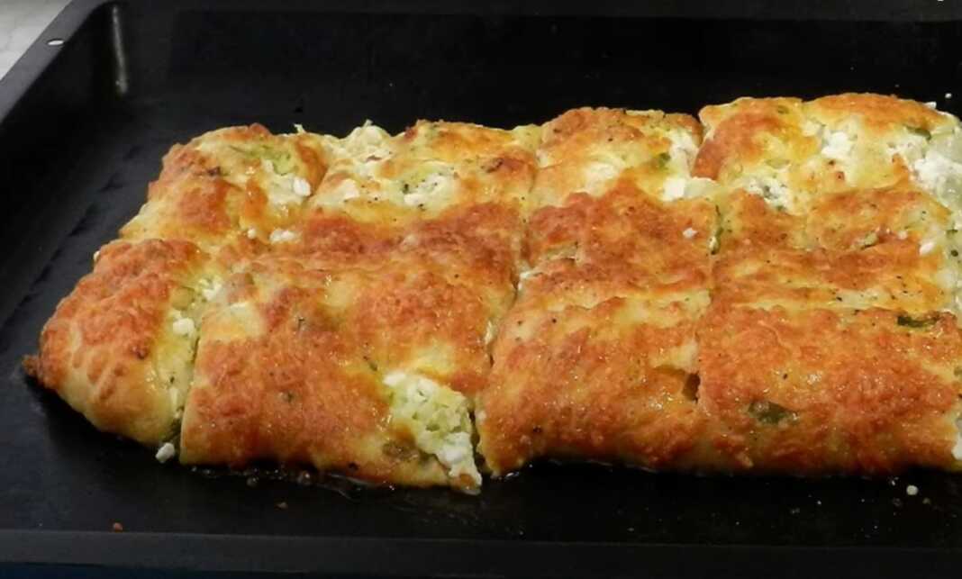 Ένα ψημένο κολοκυθοτυρόψωμο με χρυσαφένια κρούστα, κομμένο σε λωρίδες, με τριμμένο τυρί στην επιφάνεια.