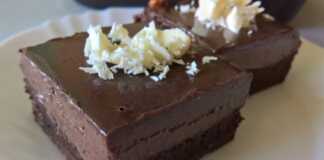 Ενα κομμάτι από μια πλούσια, σκούρα καφέ τούρτα σοκολάτας με γυαλιστερό γλάσο από πάνω, διακοσμημένη με ξύσμα λευκής σοκολάτας.