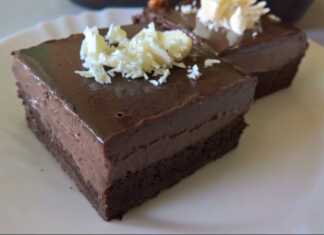 Ενα κομμάτι από μια πλούσια, σκούρα καφέ τούρτα σοκολάτας με γυαλιστερό γλάσο από πάνω, διακοσμημένη με ξύσμα λευκής σοκολάτας.