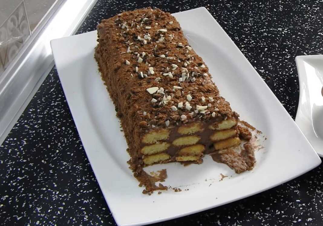 Ένας ολόκληρος σοκολατένιος κορμός με σαβαγιάρ σε μια λευκή πιατέλα, πασπαλισμένος με τριμμένη σοκολάτα ή ξηρούς καρπούς.