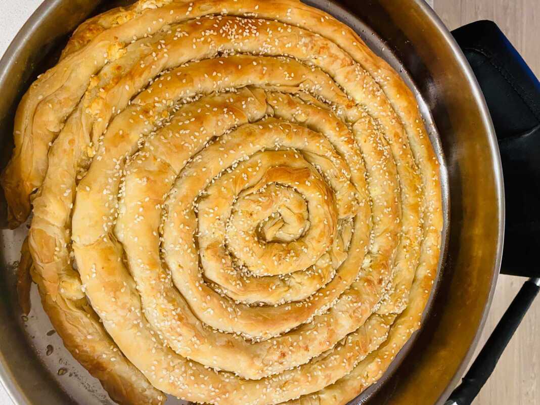 Χρυσαφένια στριφτή τυρόπιτα σε σχήμα σαλιγκαριού μέσα στο ταψί πασπαλισμένη με σουσάμι και τραγανό φύλλο