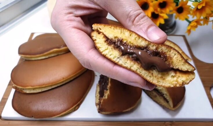 Δύο αφράτα χρυσαφένια σπιτικά pancakes ενωμένα σαν σάντουιτς, όπου από το πλάι ξεχειλίζει πλούσια ρευστή σοκολάτα Nutella πάνω σε μια λευκή πιατέλα