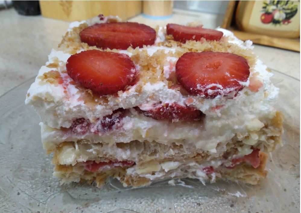 milfeigFraoules3 Ένα κομμάτι μιλφέιγ με στρώσεις χρυσής σφολιάτας, λευκής κρέμας και φρέσκων κόκκινων φραουλών, πασπαλισμένο με άχνη ζάχαρη, σε ένα λευκό πιάτο.