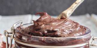 Ένα βάζο γεμάτο με λαχταριστή, καφέ, πηχτή σπιτική μερέντα, με ένα κουτάλι βουτηγμένο μέσα της και ψωμί στο φόντο.