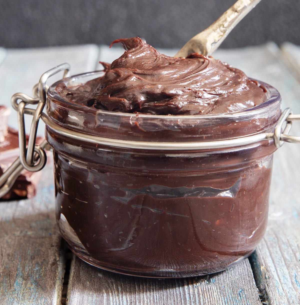 spitikiMerenta Ένα βάζο γεμάτο με λαχταριστή, καφέ, πηχτή σπιτική μερέντα, με ένα κουτάλι βουτηγμένο μέσα της και ψωμί στο φόντο.