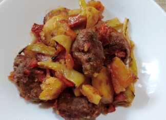 Κυλινδρικά κεφτεδάκια (σουτζουκάκια) σε ταψί, πλαισιωμένα από κομμένες πατάτες, πιπεριές και ντομάτες, όλα καλυμμένα με κόκκινη σάλτσα, έτοιμα για ψήσιμο