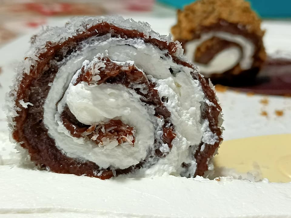 RoloSokaolatas8 Ένα ορθογώνιο ταψί με στρώσεις σοκολατένιας κρέμας και λευκής σαντιγί, πασπαλισμένο με καρύδα και καρύδι, κομμένο σε λωρίδες που ρολάρονται.