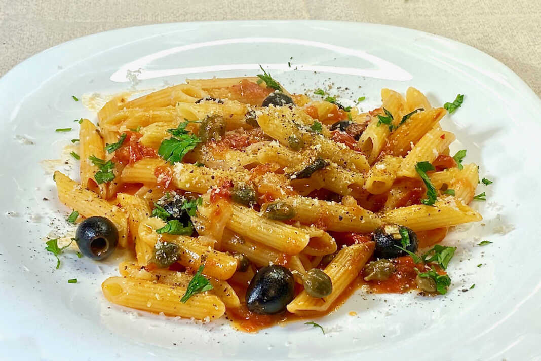 Ένα πιάτο με πένες ανακατεμένες με κόκκινη σάλτσα ντομάτας, πράσινο βασιλικό και μαύρες ελιές.