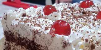 Ένα κομμάτι λευκής πάστας σε πιάτο, που δείχνει τις στρώσεις του σκούρου παντεσπανιού, της λευκής κρέμας με κομματάκια μαντολάτου και διακόσμηση με λευκή σαντιγί και ζαχαρωμένα κεράσια.