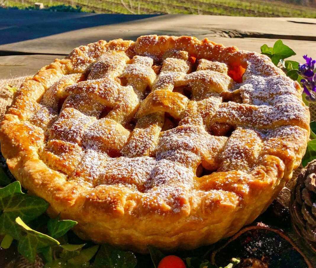 rustikMilopita Μια ολόκληρη, χρυσαφένια μηλόπιτα με πλέγμα στην κορυφή, πασπαλισμένη με άχνη ζάχαρη, πάνω σε ένα ξύλινο τραπέζι.