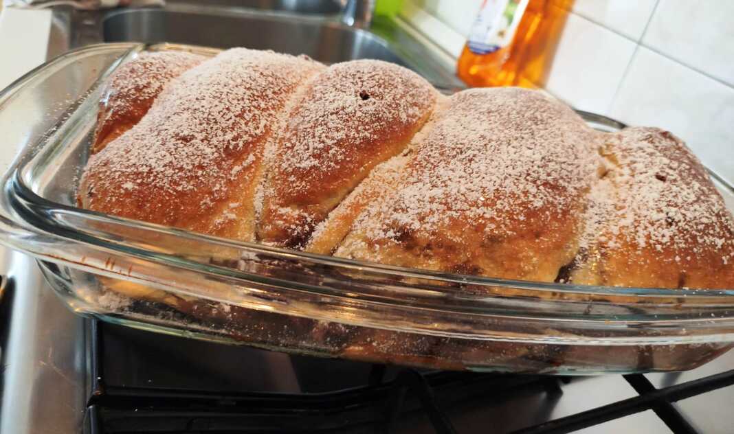 KeikKakaoKarida Ένα ολόκληρο, χρυσαφένιο κέικ πλεγμένο σε κοτσίδα, πασπαλισμένο με άχνη ζάχαρη, με τη γέμιση κακάο-καρύδας να φαίνεται διακριτικά.