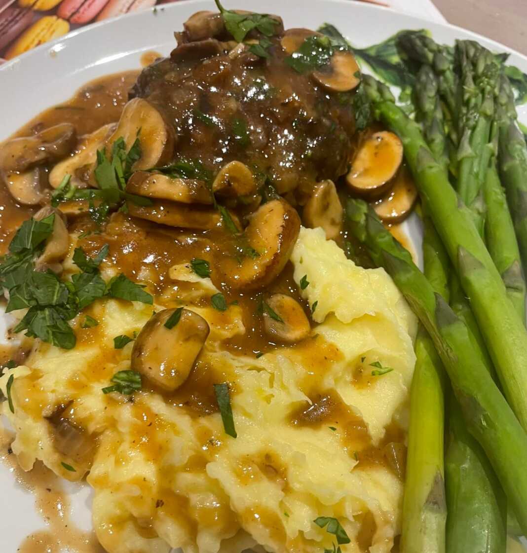Ζουμερά Μπιφτέκια σε σάλτσα μανιταριών, σερβιρισμένα με πουρέ πατάτας και πράσινα σπαράγγια, πασπαλισμένα με μαϊντανό.