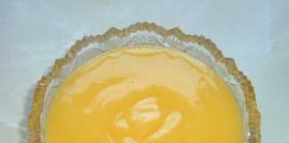 Ένα γυάλινο μπολάκι γεμάτο με λαμπερή κίτρινη κρέμα λεμονιού δίπλα σε φρεσκοκομμένα λεμόνια και μια ξύλινη κουτάλα