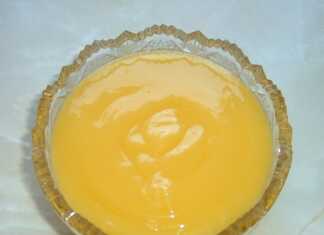 Ένα γυάλινο μπολάκι γεμάτο με λαμπερή κίτρινη κρέμα λεμονιού δίπλα σε φρεσκοκομμένα λεμόνια και μια ξύλινη κουτάλα