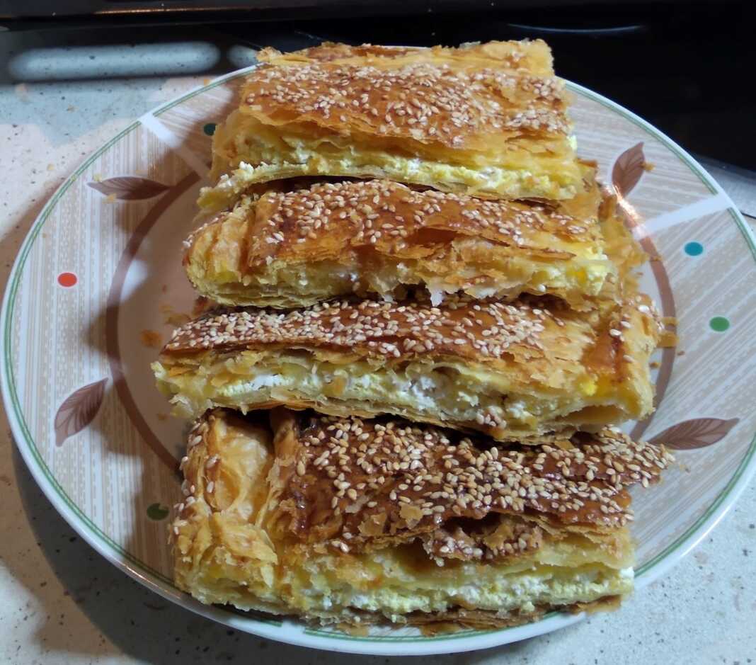 Μια χρυσαφένια, φρεσκοψημένη τυρόπιτα με σφολιάτα, κομμένη σε τετράγωνα κομμάτια, με εμφανή την τραγανή της υφή και την πλούσια γέμιση τυριού.