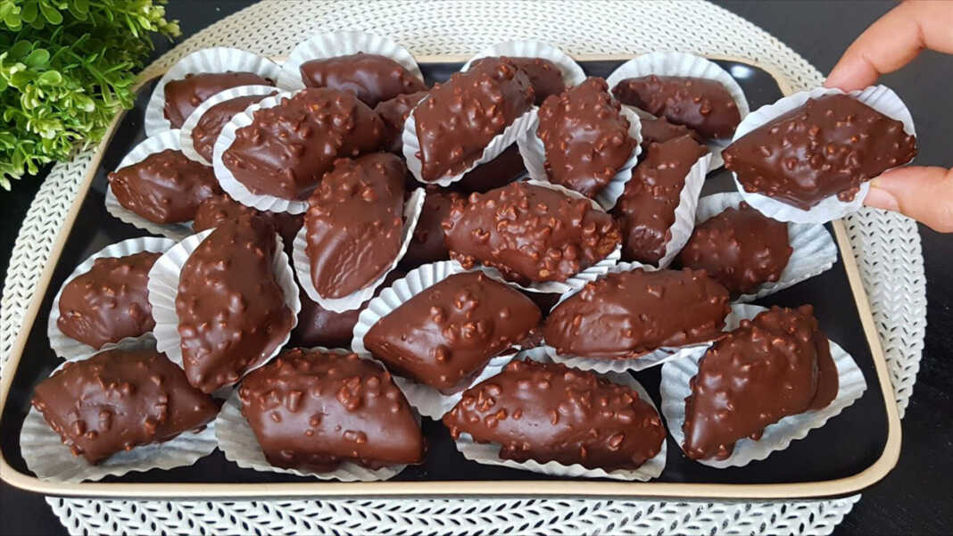 Ένα ταψί με λαδόκολλα γεμάτο με σκουρόχρωμα, σοκολατένια μπισκότα, μερικά από τα οποία είναι ήδη βουτηγμένα σε λιωμένη σοκολάτα και πασπαλισμένα με αμύγδαλα.