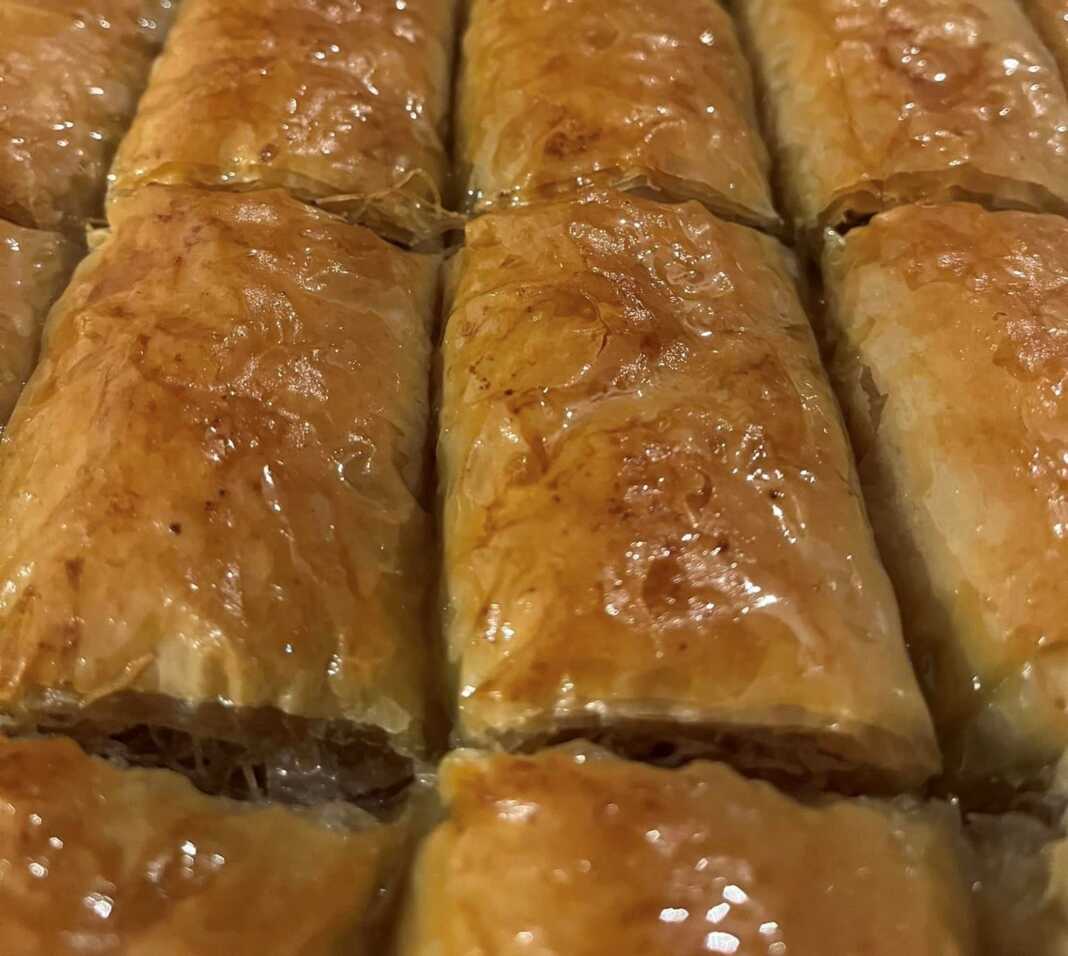 Ένα ταψί με χρυσαφένια ρολά κανταΐφι, κομμένα σε μερίδες, με χοντροκομμένα καρύδια και αμύγδαλα στην επιφάνεια.