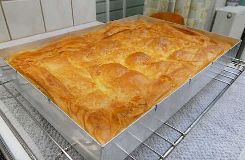 mpougatsaThessaloniki Μια φρεσκοψημένη, χρυσαφένια μπουγάτσα Θεσσαλονίκης, κομμένη σε τετράγωνα κομμάτια, πασπαλισμένη με ζάχαρη άχνη και κανέλα.