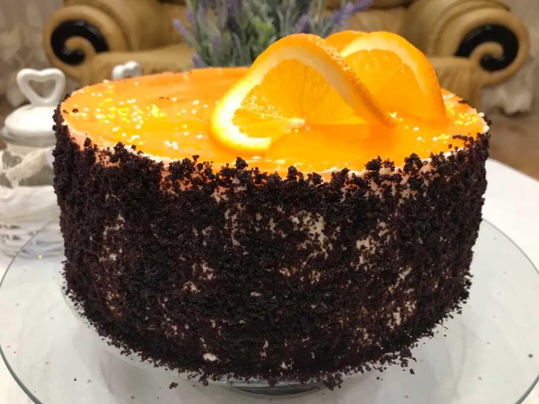 Ένα Κεικ πολύστρωτο με βάση σοκολάτας, ενδιάμεσες στρώσεις λευκής κρέμας τυριού και πορτοκαλί γλάσου, και ψίχουλα κέικ στα πλαϊνά.