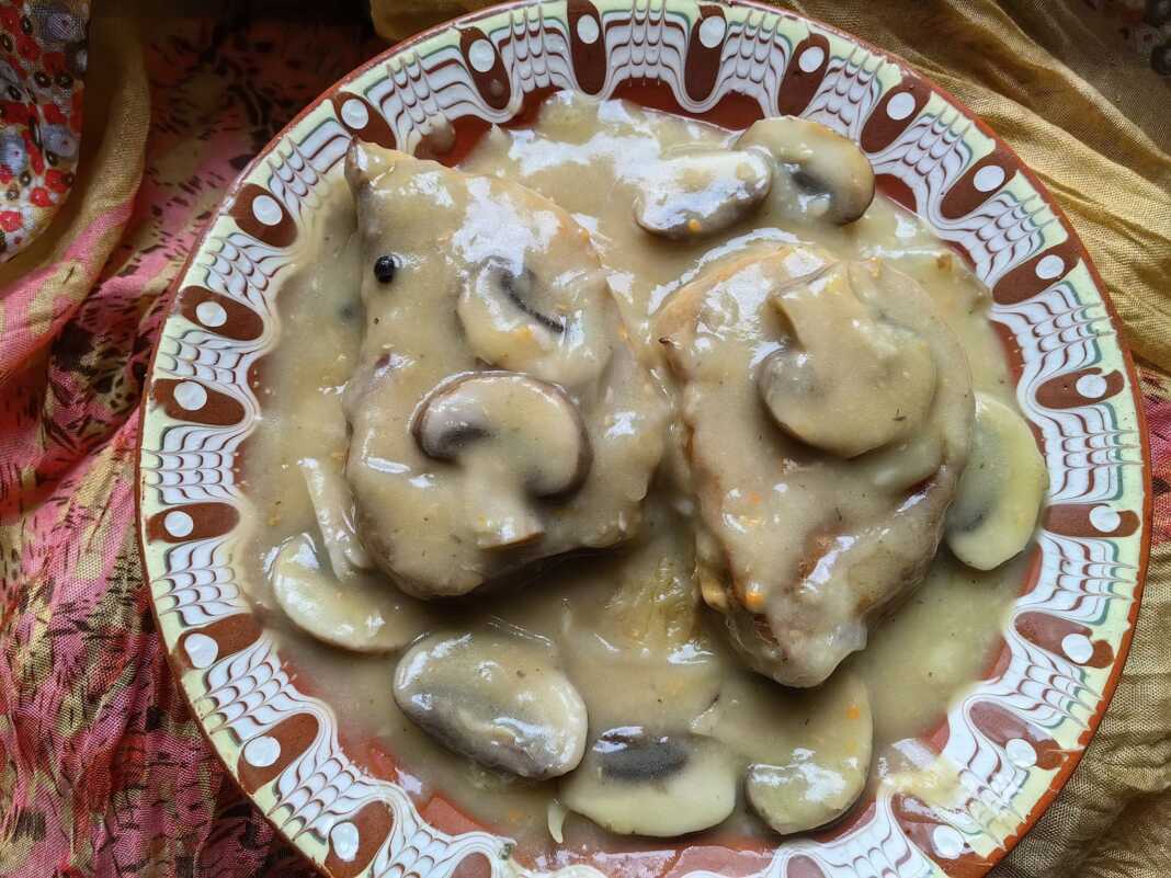 Δύο χρυσαφένιες χοιρινές μπριζόλες με πλούσια καφέ σάλτσα μανιταριών και φρέσκα μυρωδικά σε ένα λευκό πιάτο.