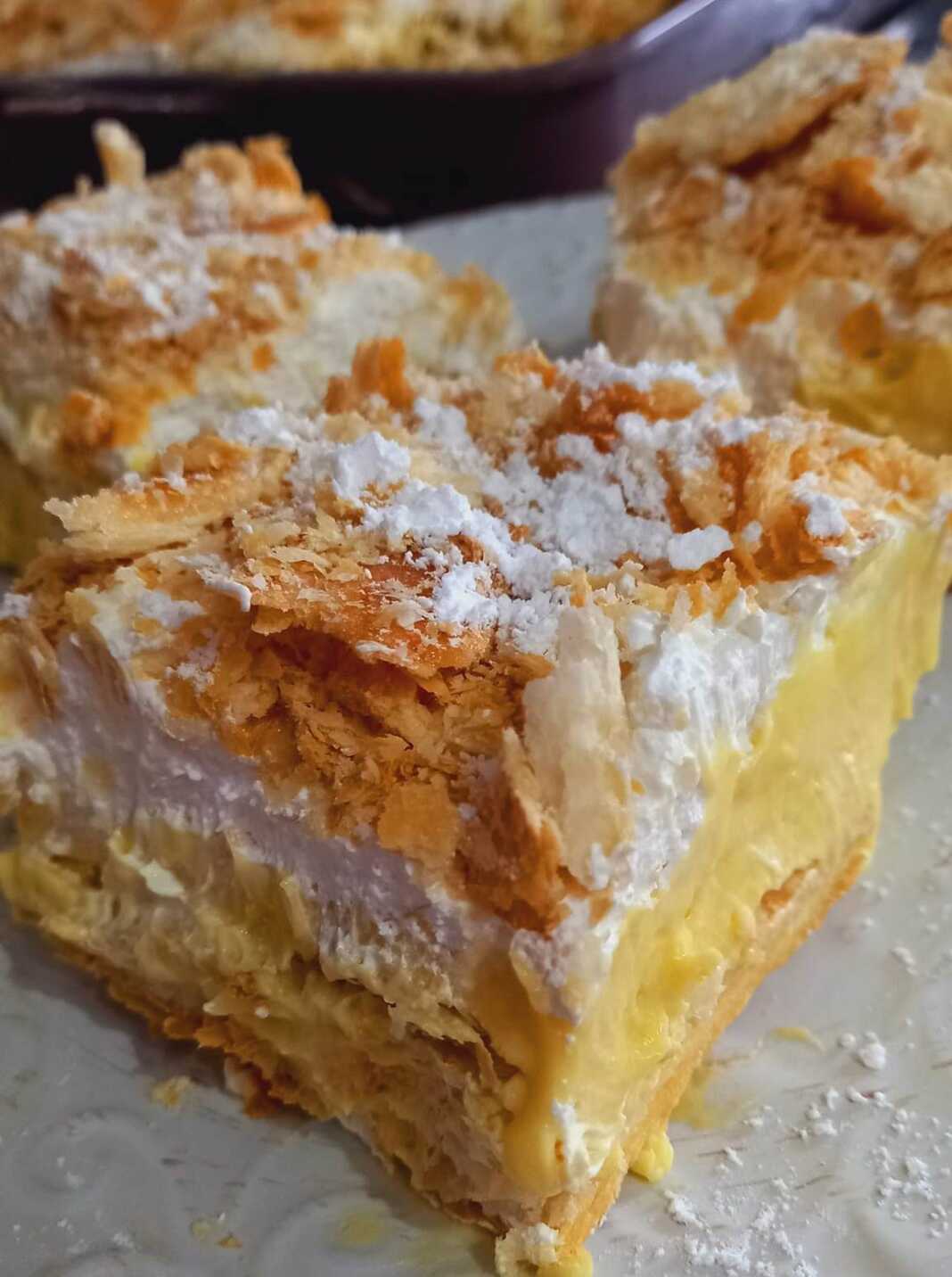 Ένα κομμάτι μιλφέιγ με εμφανή τα τραγανά φύλλα σφολιάτας και την κρεμώδη γέμιση, πασπαλισμένο με άχνη ζάχαρη.