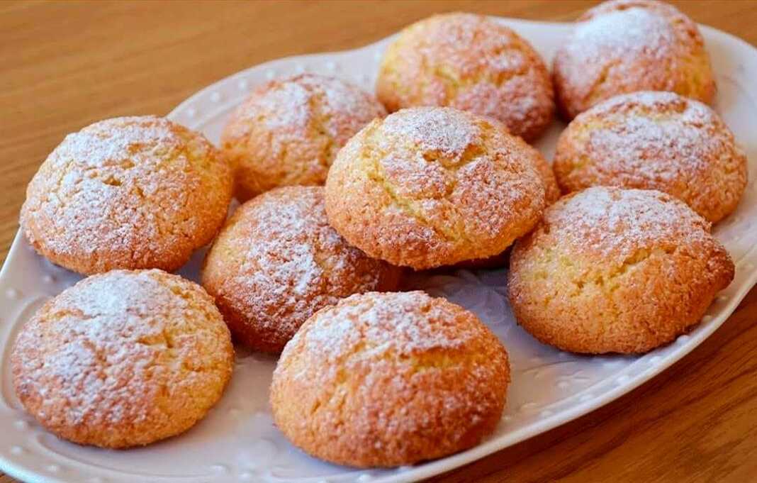 mpiskotaKaridas Ένα πιάτο γεμάτο με χρυσαφένια, στρογγυλά μπισκότα καρύδας, πασπαλισμένα με ζάχαρη άχνη, έτοιμα για σερβίρισμα.