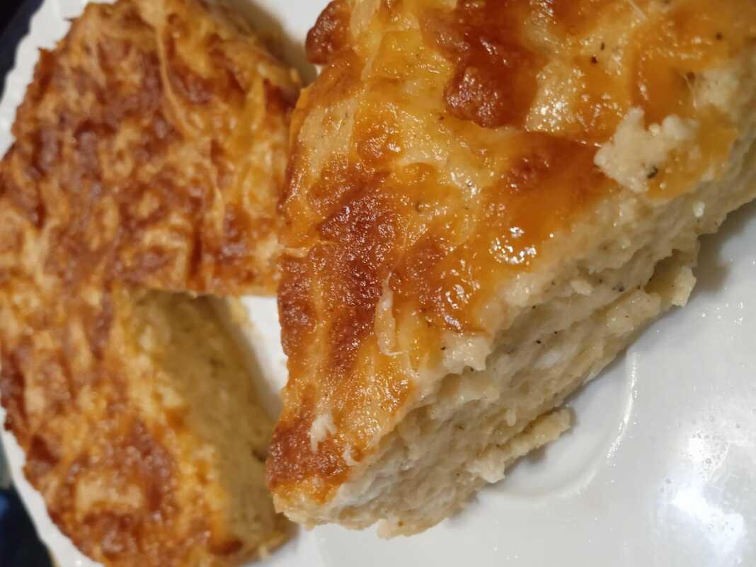 Ένα κομμάτι χρυσαφένιας κασερόπιτας χωρίς φύλλο σε ένα λευκό πιάτο. Η πίτα φαίνεται αφράτη και έχει τριμμένο κασέρι στην επιφάνεια.