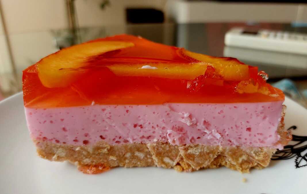 cheesecakeZele Ένα κομμάτι δροσερού cheesecake με μπισκοτένια βάση, λευκή κρέμα γιαουρτιού και δύο στρώσεις χρωματιστού ζελέ, γαρνιρισμένο με φέτες νεκταρινιού, σερβιρισμένο σε λευκό πιάτο.