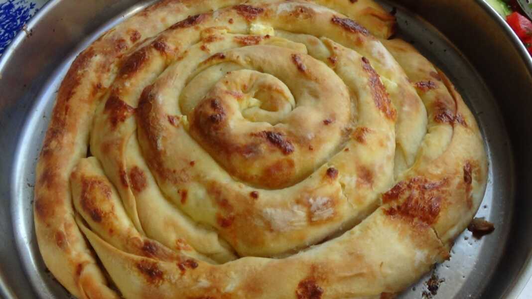 Στρογγυλή, χρυσαφένια ζυμωτή πίτα τοποθετημένη κυκλικά σε ταψί, γεμισμένη με φέτα και γιαούρτι, έτοιμη να κοπεί.