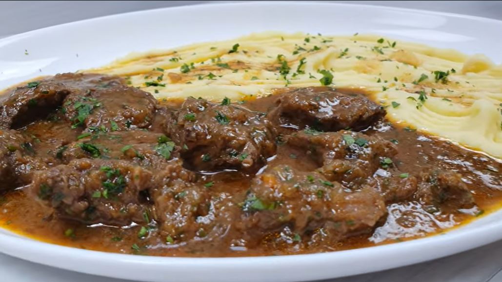 mosxarakiKatsarolas Ένα πιάτο με κομμάτια μοσχαρίσιου κρέατος με καφεκόκκινη σάλτσα, δίπλα σε έναν λευκό αφράτο πουρέ πατάτας, γαρνιρισμένο με φρέσκο μαϊντανό.