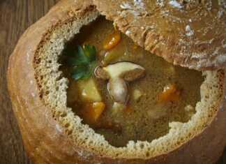 Αχνιστή σούπα με κρέας και λαχανικά μέσα σε ένα σκαμμένο καρβέλι μαύρο ψωμί με το καπάκι του στο πλάι και φρέσκα μυρωδικά