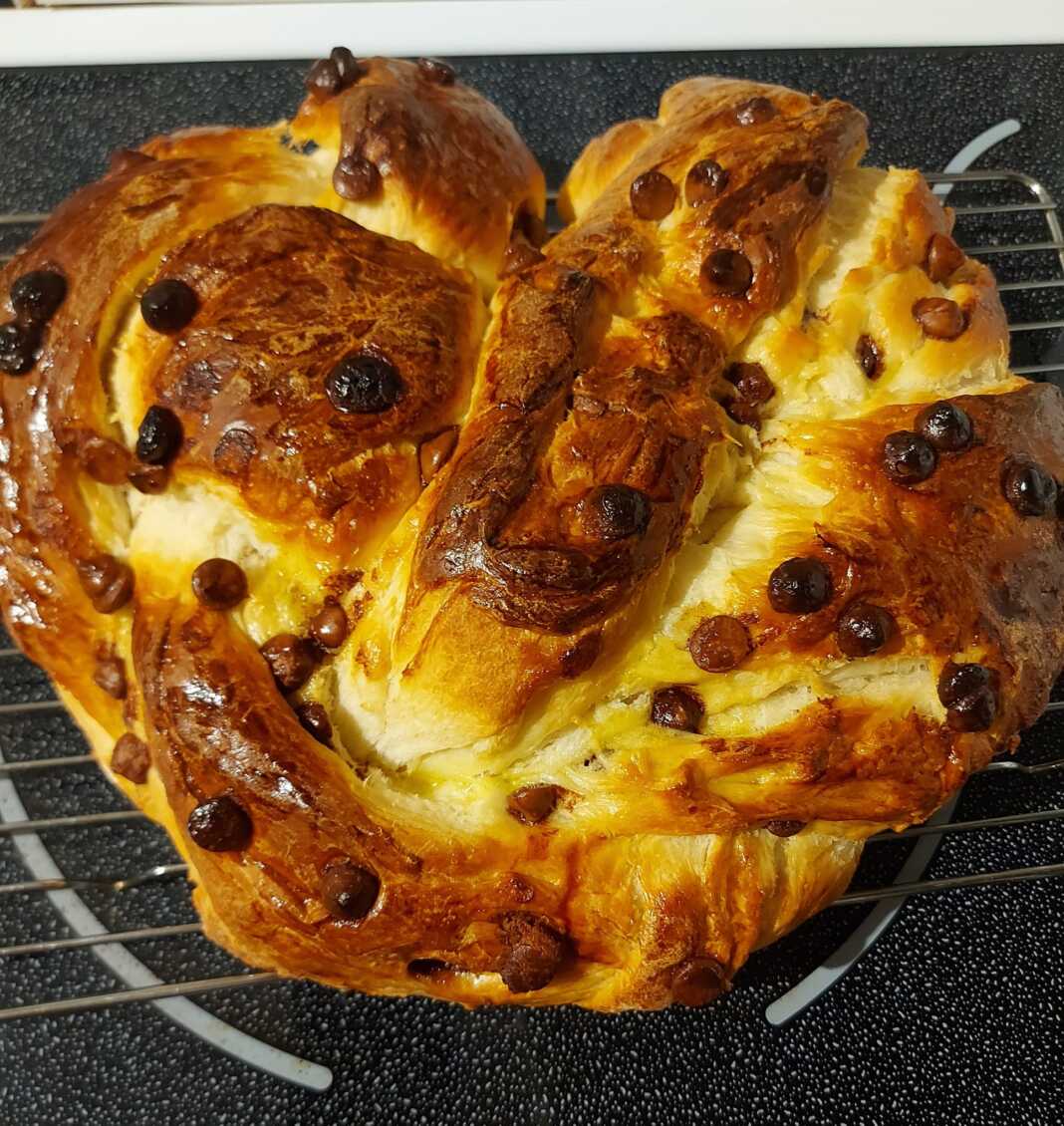 Ένα αφράτο, χρυσαφένιο τσουρέκι πλεγμένο σε πλεξούδα, με κομματάκια σοκολάτας στην επιφάνεια, σε ένα στρογγυλό ταψί με λαδόκολλα.