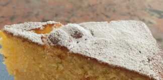 Ένα ολόκληρο, στρογγυλό κέικ αμυγδάλου πασπαλισμένο με ζάχαρη άχνη, τοποθετημένο σε λευκή βάση με μπλε λουλούδια.