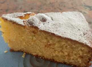 Ένα ολόκληρο, στρογγυλό κέικ αμυγδάλου πασπαλισμένο με ζάχαρη άχνη, τοποθετημένο σε λευκή βάση με μπλε λουλούδια.