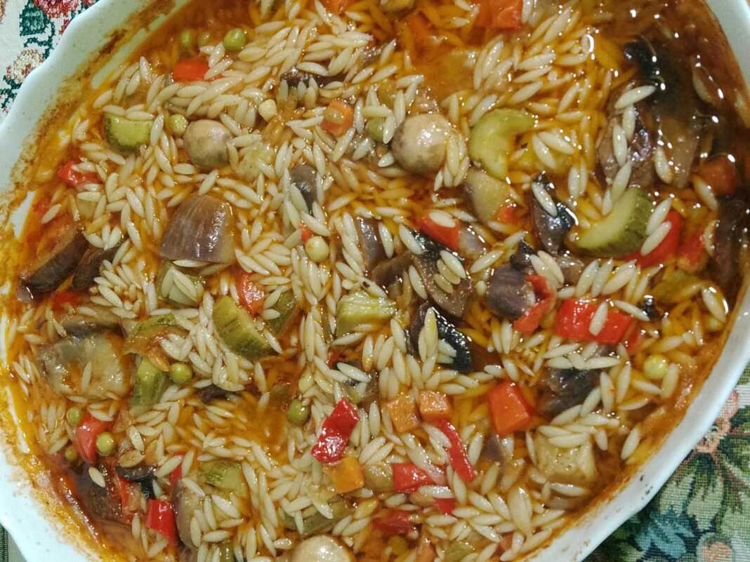 Ένα ταψί με γιουβέτσι λαχανικών, όπου διακρίνονται το κριθαράκι και τα πολύχρωμα λαχανικά σε κόκκινη σάλτσα.