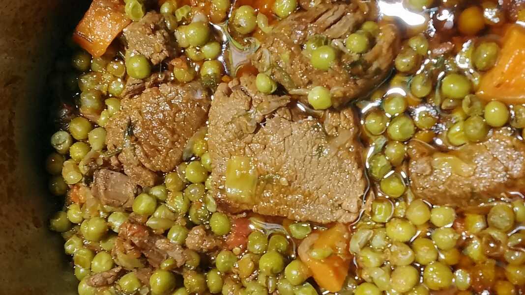 Μία κατσαρολα με μοσχαράκι κοκκινιστό, πράσινο αρακά και πορτοκαλί καρότα σερβιρισμένο σε βαθύ πιάτο.