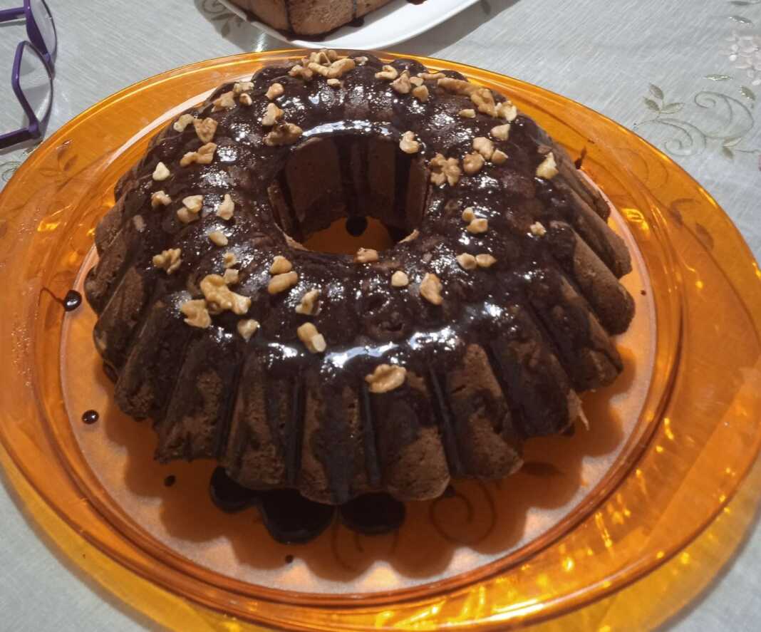 Keik nistisimo_oty Ένα νηστίσιμο κέικ πορτοκαλιού με γλάσο σοκολάτας και πασπαλισμένα καρύδια σε μια πιατέλα.