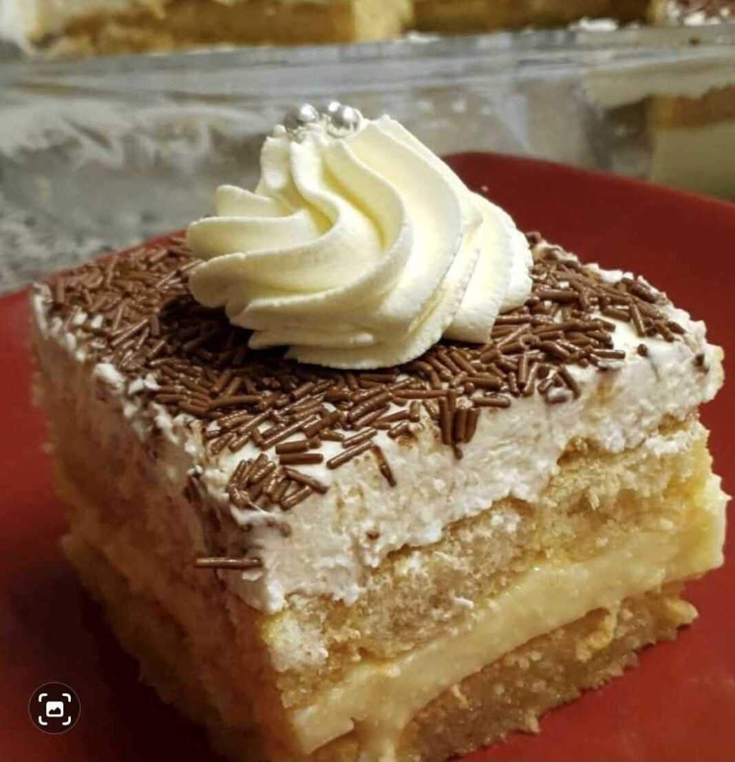 Ένα κομμάτι γλυκού σε ένα πιάτο, με στρώσεις από σιροπιασμένες φρυγανιές, μια παχιά στρώση κίτρινης κρέμας και λευκή σαντιγί στην κορυφή, πασπαλισμένο με κανέλα.