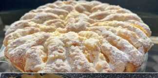 Μια στρογγυλή, χρυσαφένια τούρτα με φέτες ροδάκινου τοποθετημένες σε σχέδιο και μια στρώση ψίχουλων στην επιφάνεια, σε μια λευκή πιατέλα.