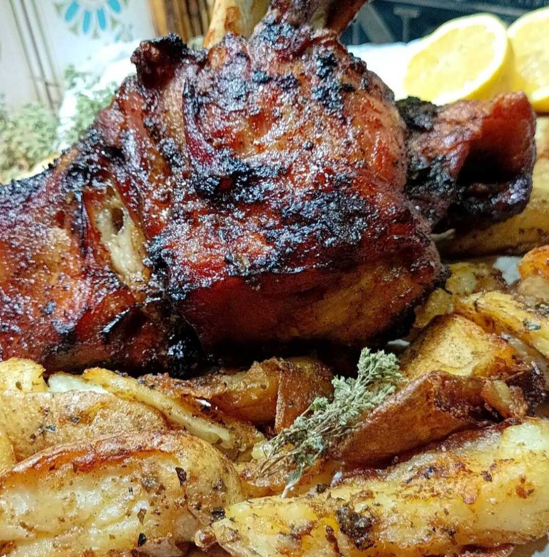 Ένα χοιρινό κότσι ψημένο στο φούρνο με σκούρα μπύρα, με τραγανή καφέ πετσούλα, σε ένα βαθύ σκεύος. Δίπλα διακρίνονται ψημένες, σπασμένες πατάτες.