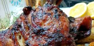 Ένα χοιρινό κότσι ψημένο στο φούρνο με σκούρα μπύρα, με τραγανή καφέ πετσούλα, σε ένα βαθύ σκεύος. Δίπλα διακρίνονται ψημένες, σπασμένες πατάτες.