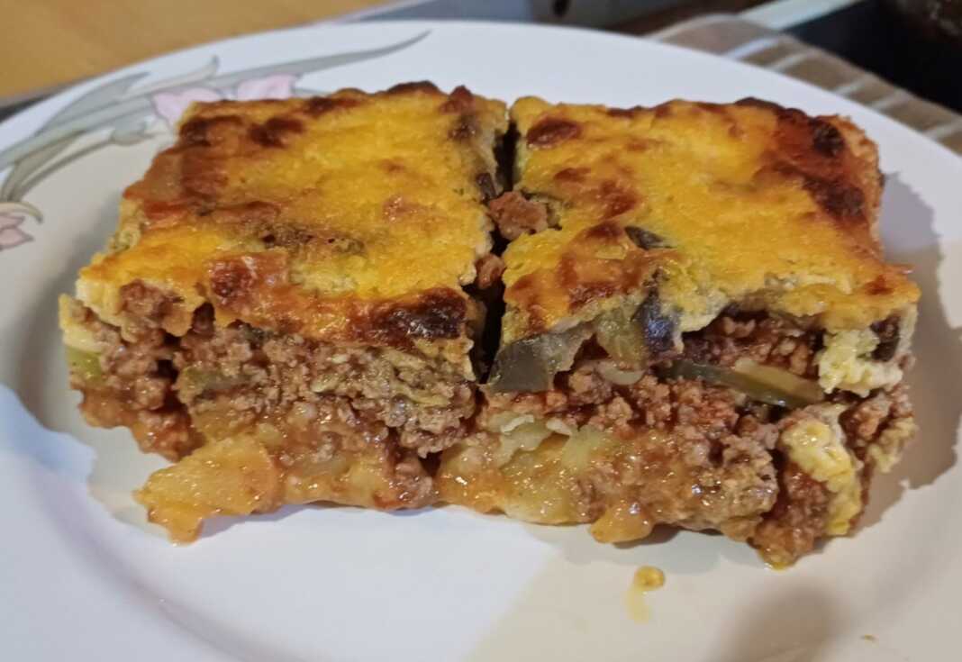 Δύο κομμάτια χρυσαφένιο μουσακά, όπου διακρίνονται οι στρώσεις λαχανικών και κιμά, με μια παχιά κρούστα μπεσαμέλ από πάνω.