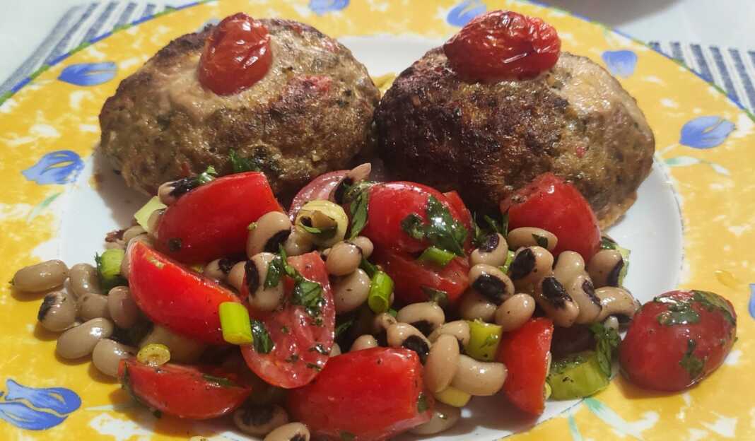 Δύο μπιφτέκια γεμιστά με τυρί σε ένα ταψί με λαδόκολλα, δίπλα σε ένα μπολ με σαλάτα από μαυρομάτικα φασόλια και κόκκινα ντοματίνια.