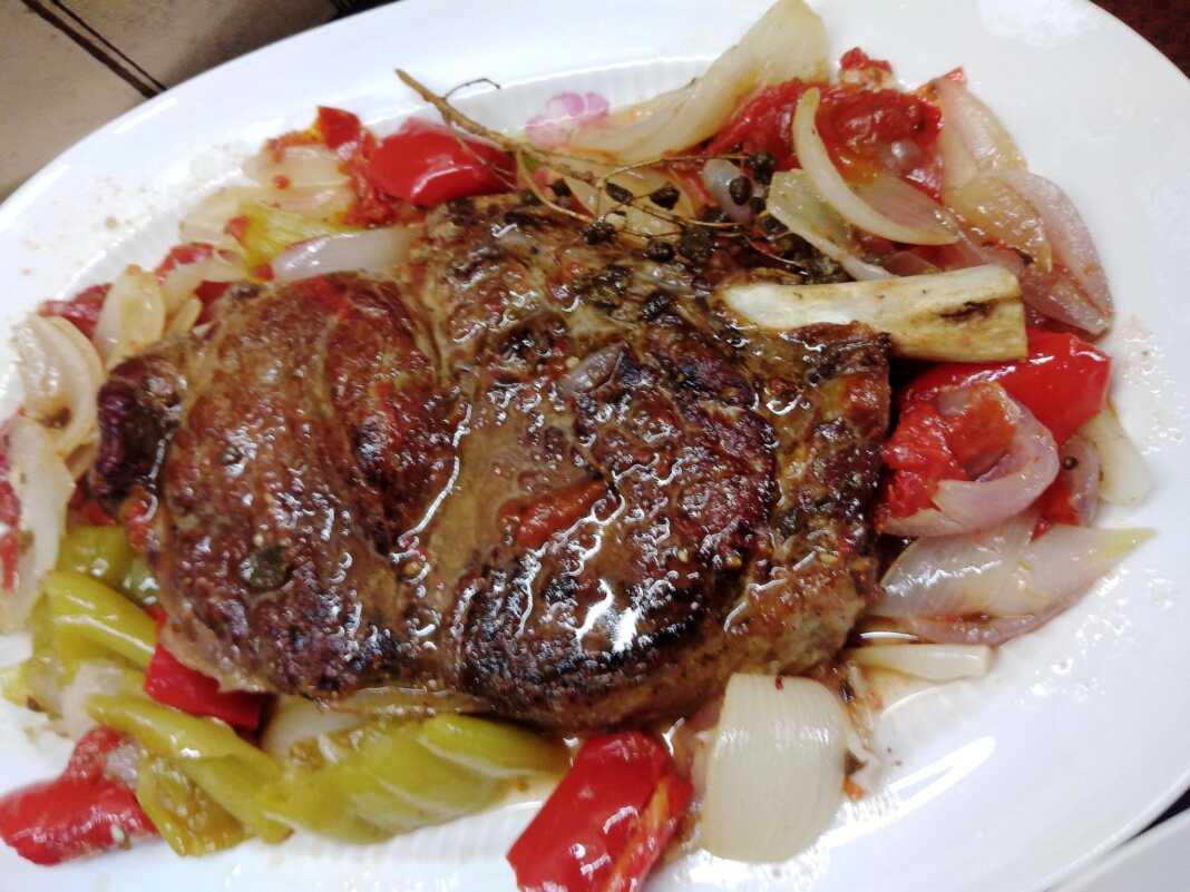 Mprizola stin ladokola Μια ψημένη μοσχαρίσια μπριζόλα σε ένα ανοιχτό πακέτο λαδόκολλας, περιτριγυρισμένη από μαγειρεμένα κομμάτια ντομάτας, κρεμμυδιού και πιπεριάς.