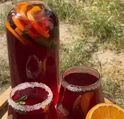 Δυο ποτήρια γεμάταμε κόκκινο δροσιστικό ρόφημα, παγάκια, φέτες πορτοκαλιού και λεμονιού, και κλωναράκια μέντας.