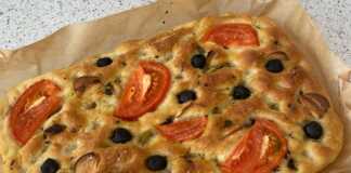 Μια ψημένη, χρυσαφένια ιταλική φοκάτσια με ντοματίνια, ελιές και φύλλα βασιλικού στην επιφάνεια, σε ένα ταψί με λαδόκολλα.