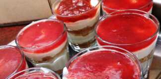 7 γυάλινα μπολακια με στρώσεις από τριμμένα μπισκότα, λευκή κρέμα γιαουρτιού και κόκκινη μαρμελάδα φράουλα στην κορυφή.