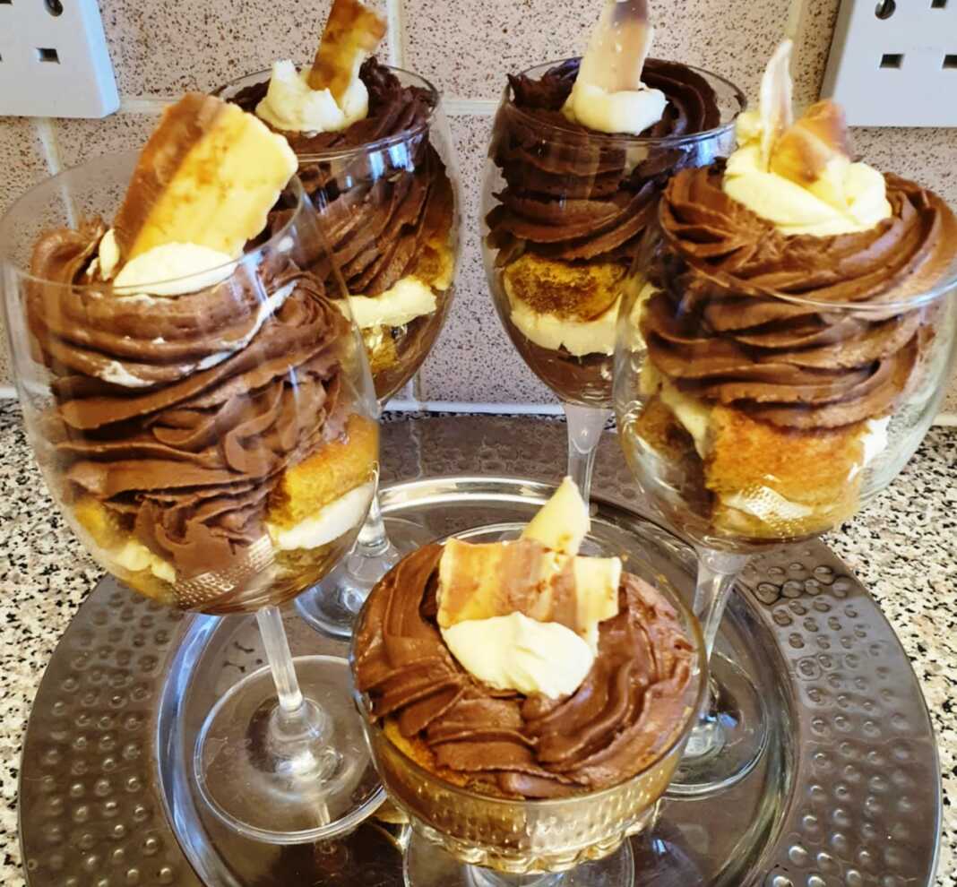 Πέντε γυάλινα ποτήρια με στρώσεις από βρεγμένα μπισκότα, άσπρη σαντιγί και σοκολατένια σαντιγί, στολισμένα με ροζ λουλούδια ζαχαρόπαστας.