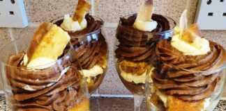 Εύκολο Γλυκάκι στο Ποτήρι Πέντε γυάλινα ποτήρια με στρώσεις από βρεγμένα μπισκότα, άσπρη σαντιγί και σοκολατένια σαντιγί, στολισμένα με ροζ λουλούδια ζαχαρόπαστας.